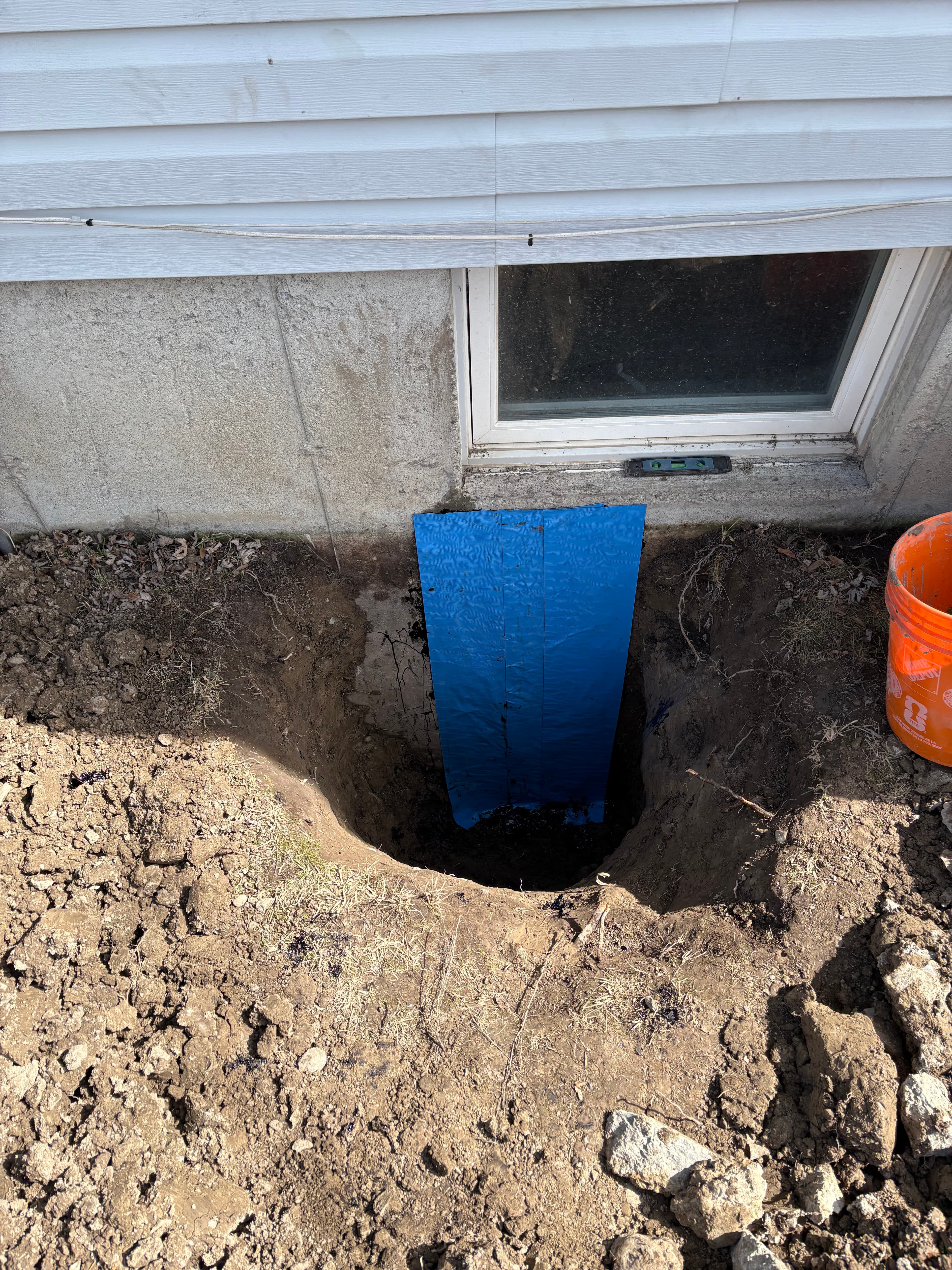 Deep excavation beside a concrete foundation wall with blue waterproofing membrane and a basement window.