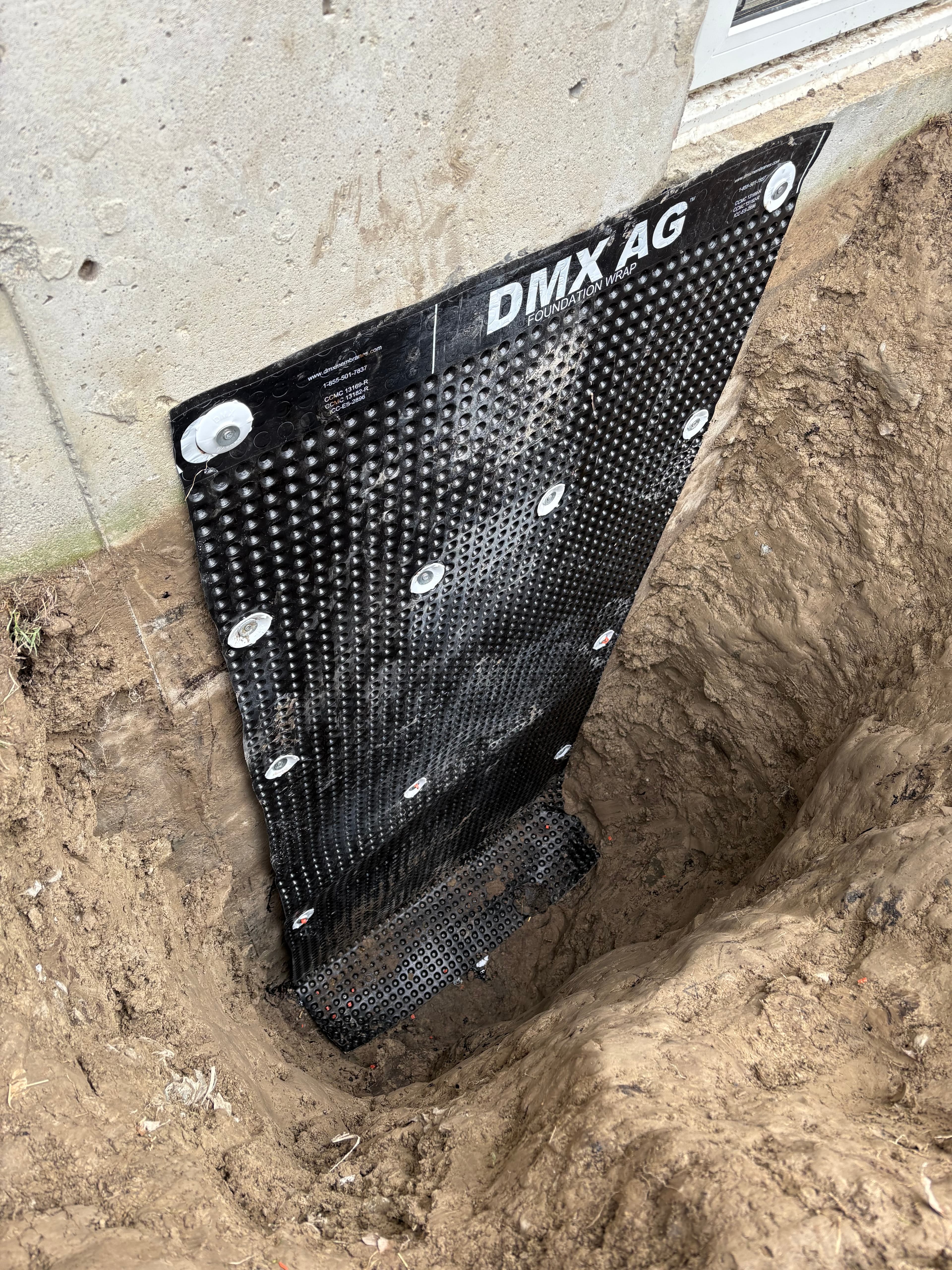 Black dimpled foundation wrap secured to a concrete wall within a deep dirt trench.