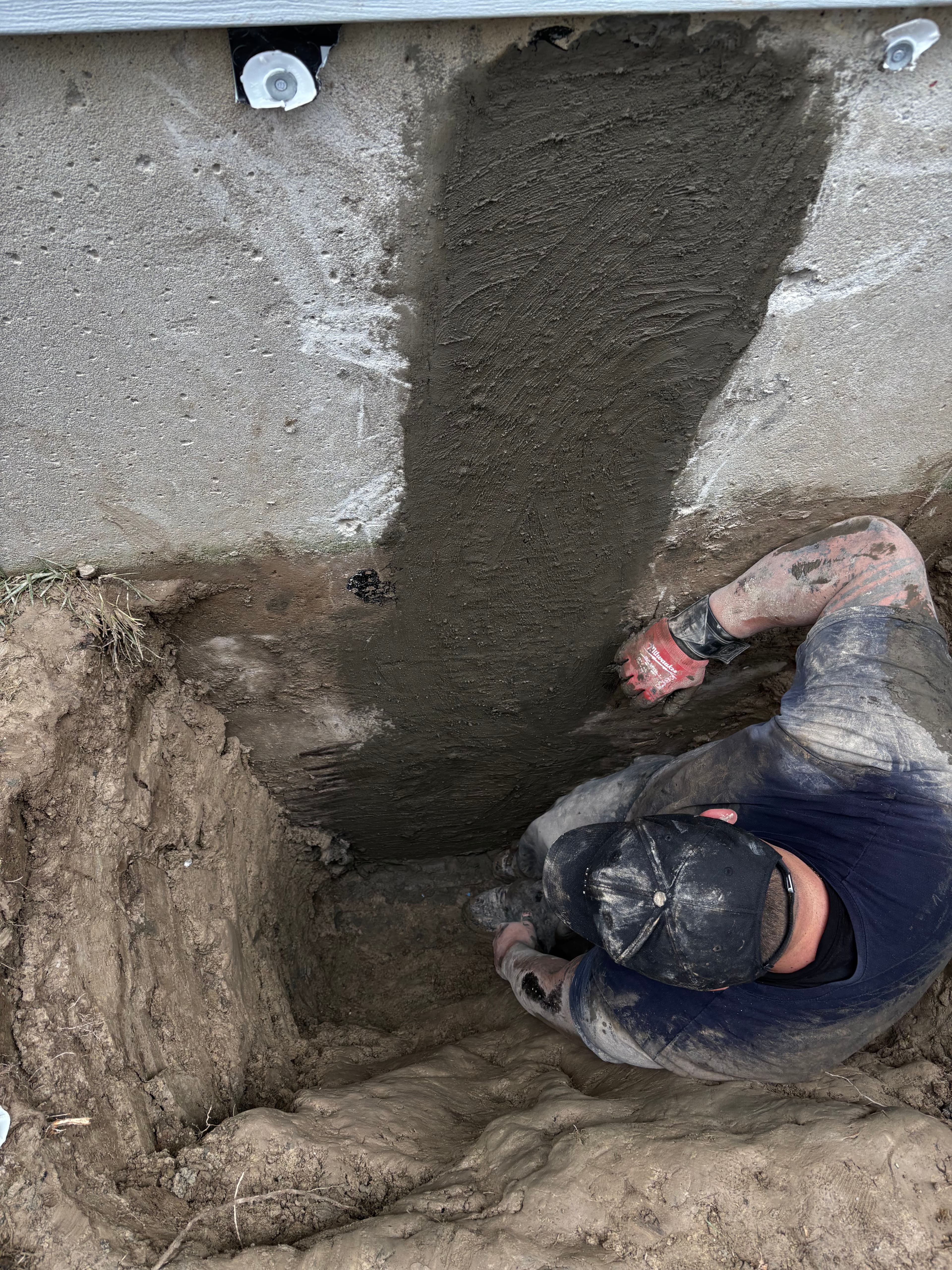Mud-covered worker in a trench applying dark sealant to a concrete foundation wall.