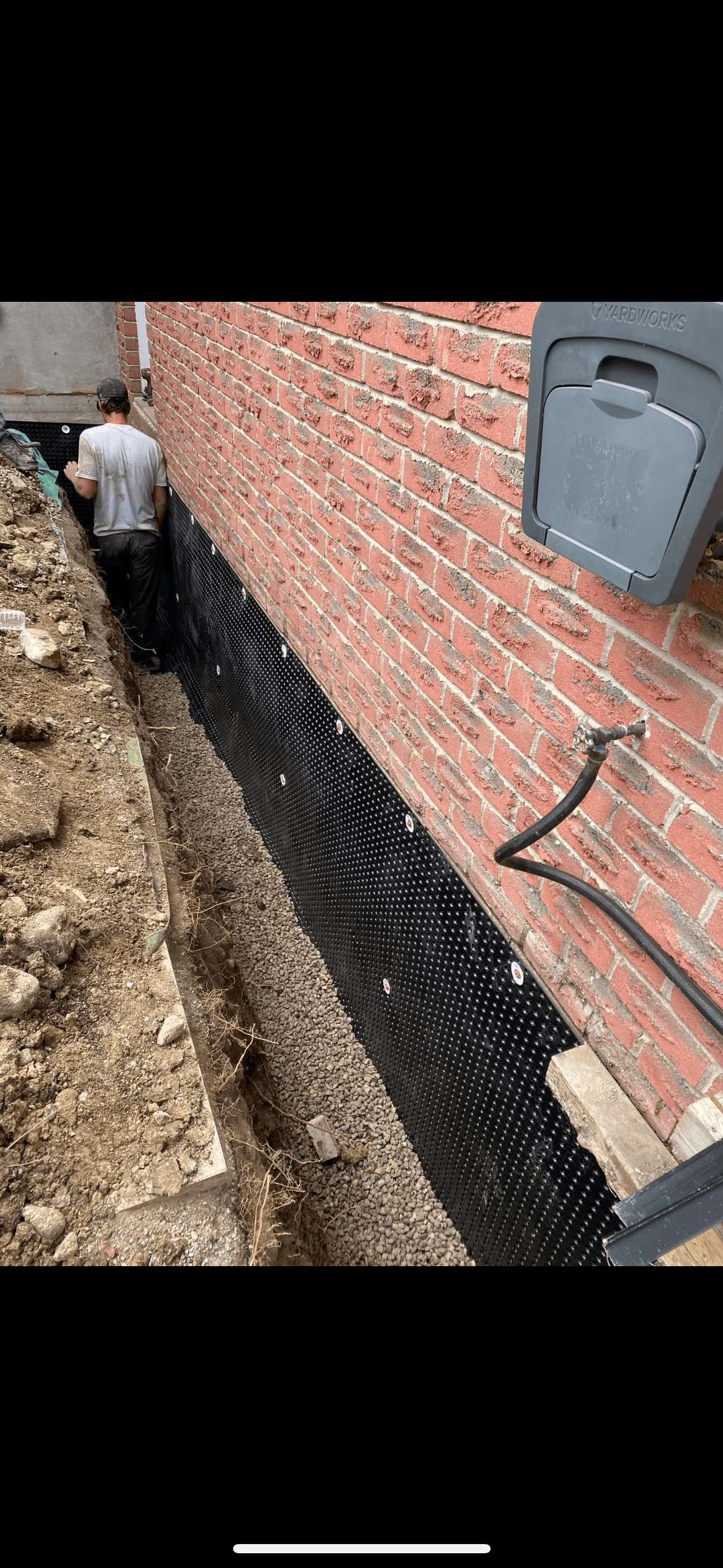 Worker installs black dimpled membrane on a red brick foundation wall in a gravel trench.