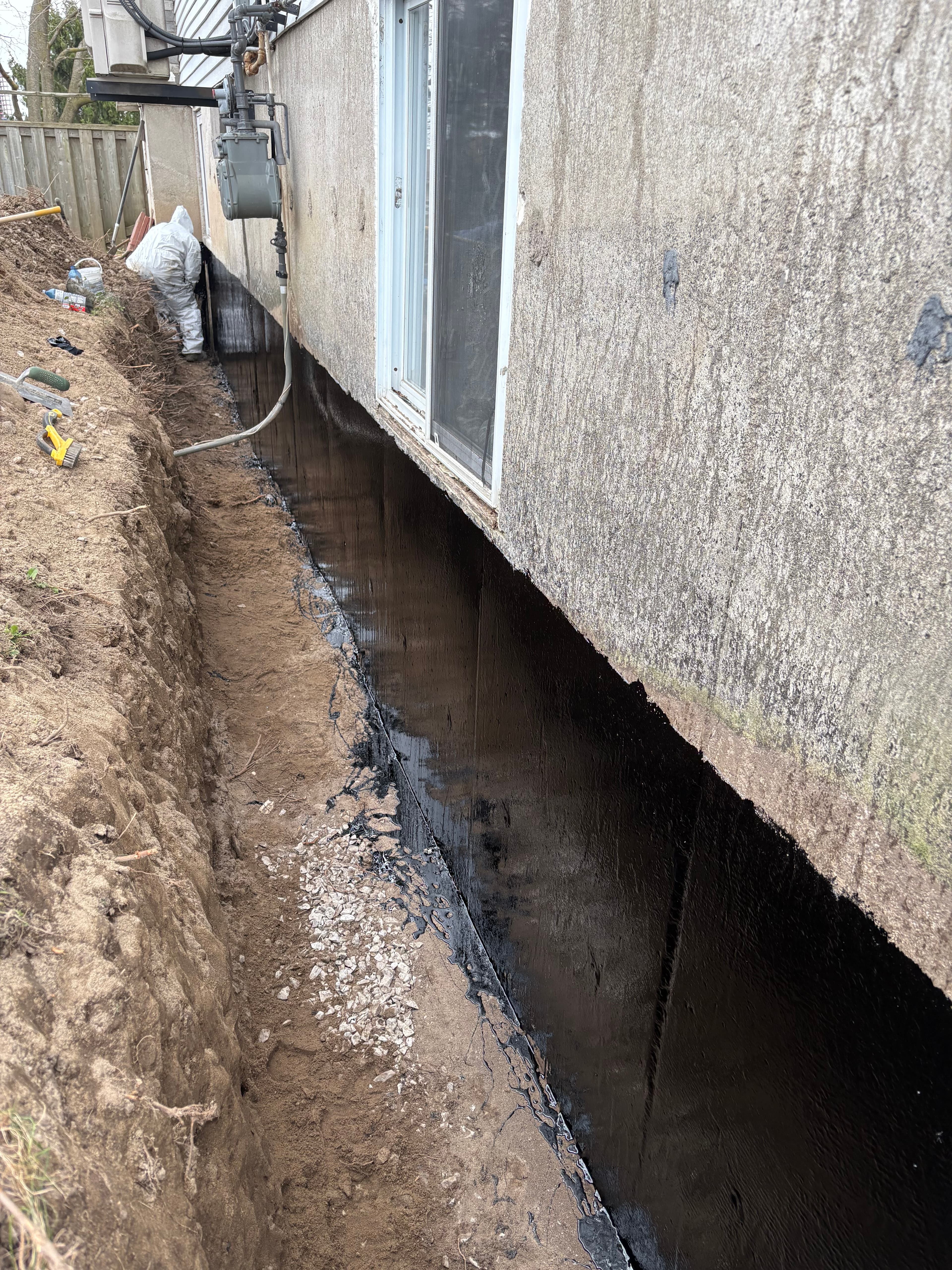 Black waterproofing sealant applied to a house foundation wall within a deep dirt trench.