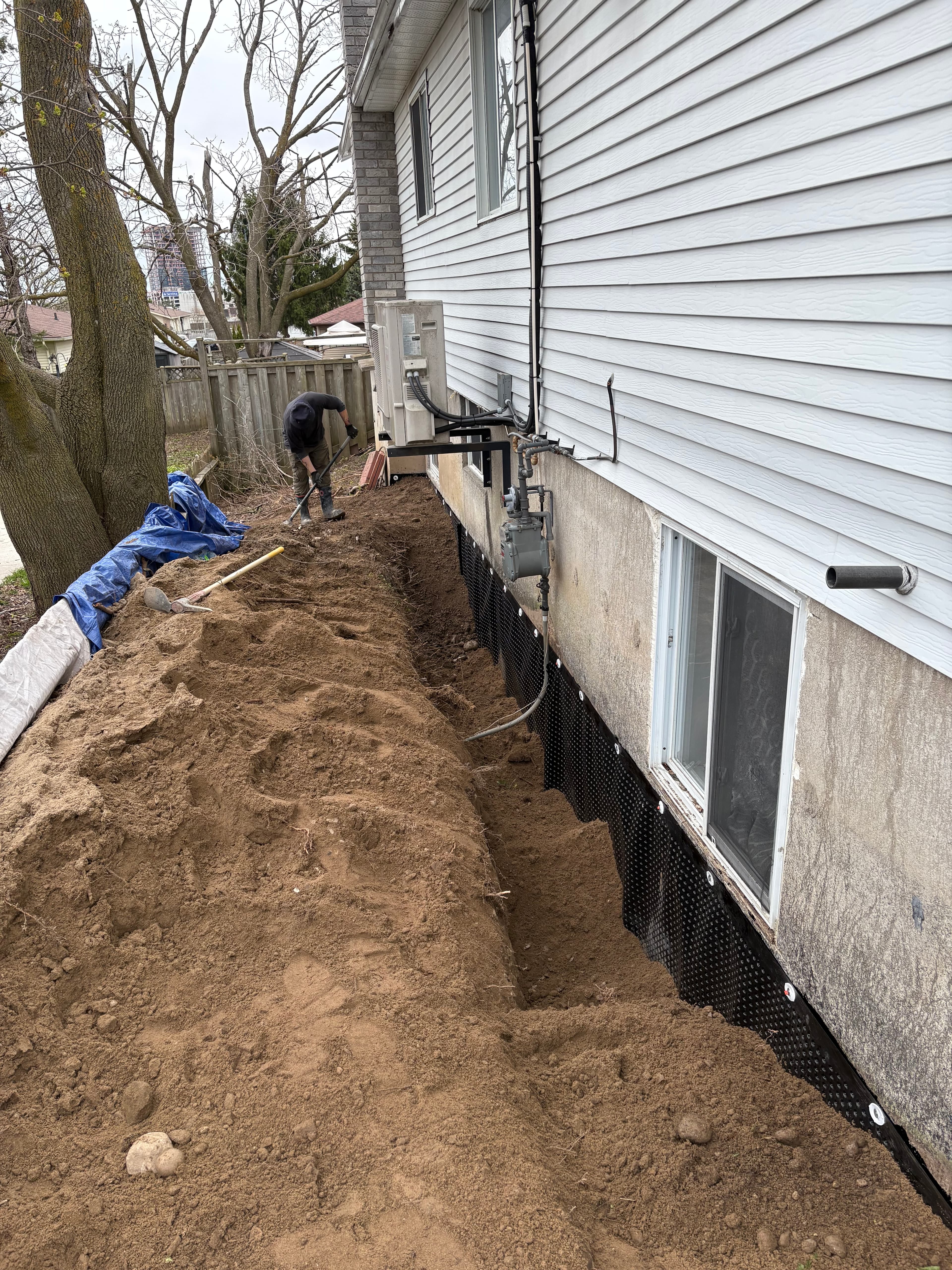 Worker digs a trench along a house foundation covered with black dimpled waterproofing membrane.