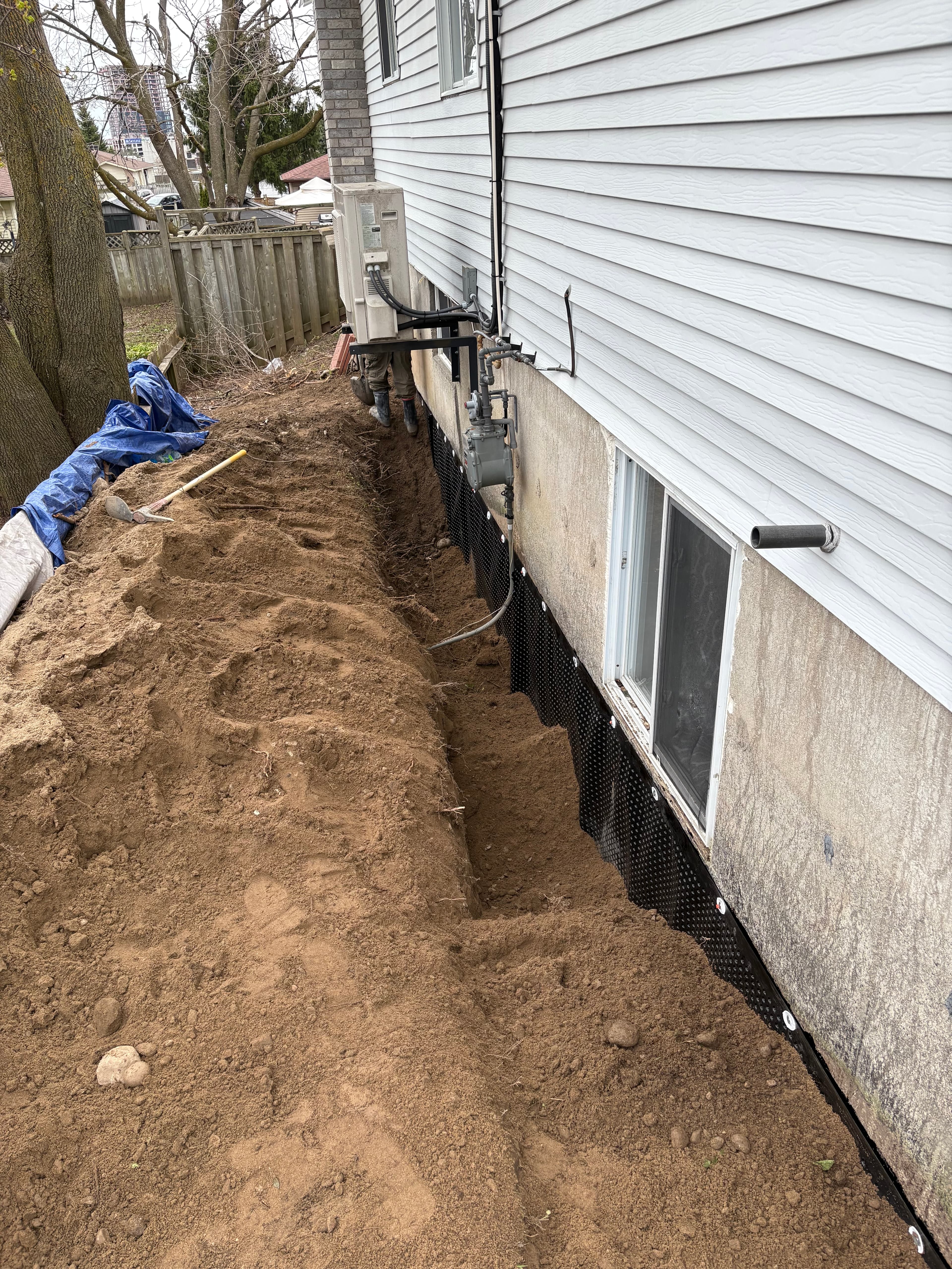 Excavated trench along a house foundation wall with black dimpled waterproofing membrane and loose soil.