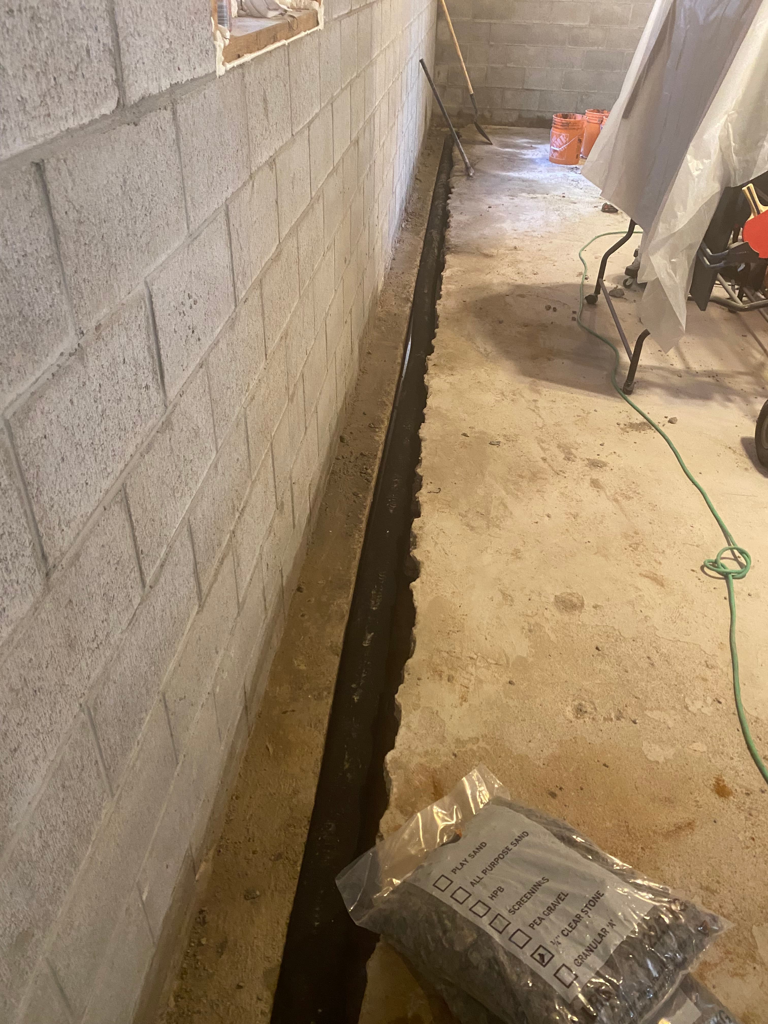 Black drainage pipe in a floor trench along a grey cinder block basement wall.