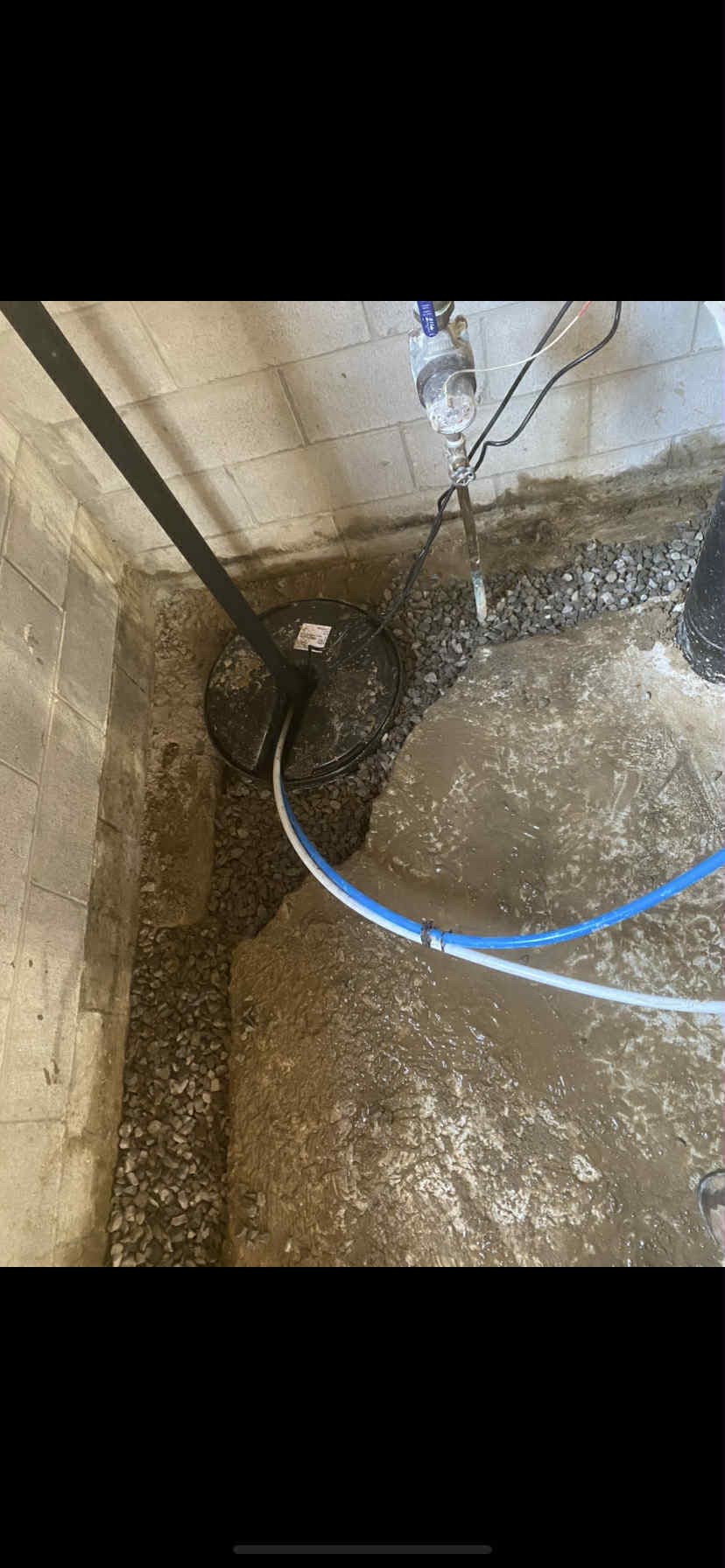 Sump pump basin in a basement corner surrounded by gravel and freshly poured concrete.